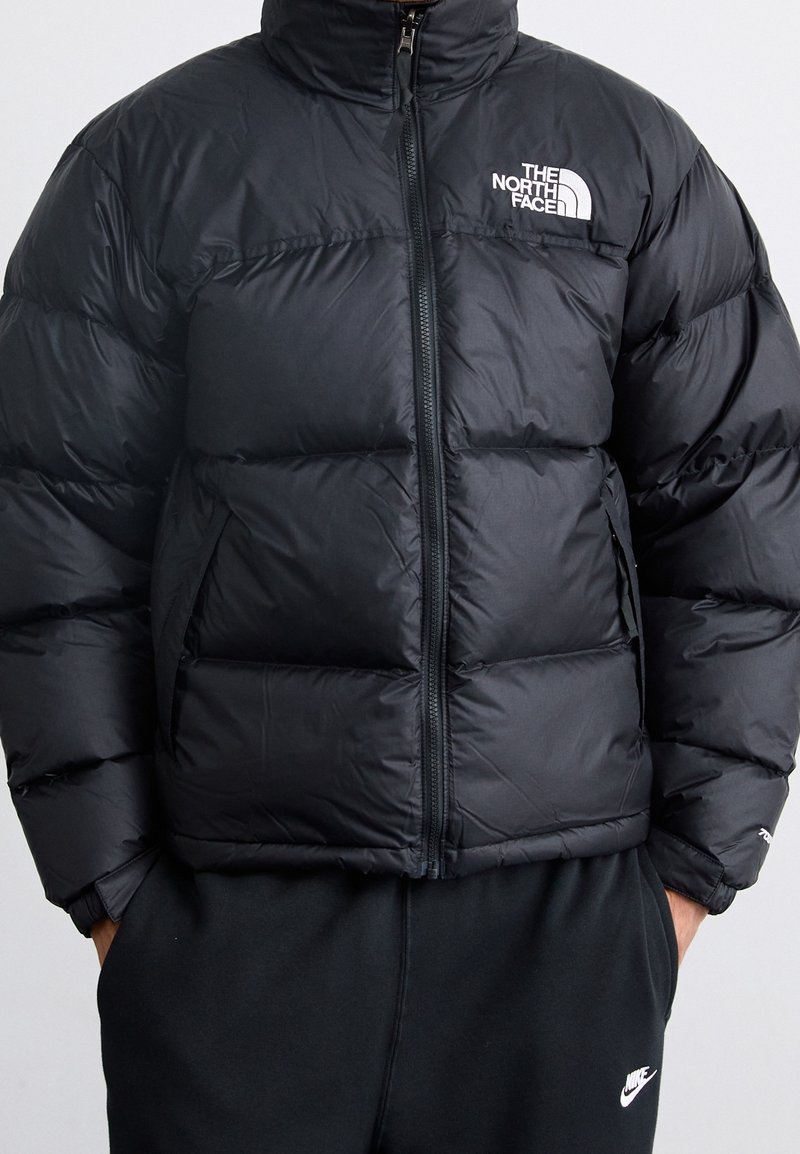 Doudoune noire avec fermeture éclair à l'avant et logo The North Face portée par une personne avec les mains dans les poches d'un pantalon de survêtement Nike noir.
