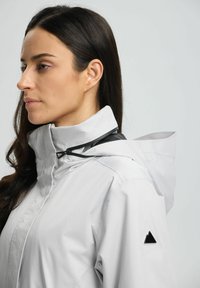 Chaqueta impermeable de color gris claro con un alto cuello y capucha ajustable. Presenta herrajes negros y un pequeño logo triangular en el hombro.