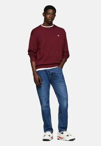Suéter de punto en color marrón, con puños acanalados, sobre una camiseta blanca de cuello redondo, combinado con jeans azules y zapatillas blancas con acentos rojos.