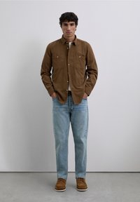 Bruin overhemd met knopen en twee borstzakken, lichtblauwe spijkerbroek en beige schoenen. Neutrale achtergrond met een subtiele textuur. Simpele, casual outfit.