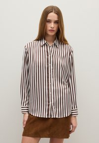 Young woman wearing a brown and white vertical striped button-up blouse and a brown suede mini skirt, standing against a plain wall.