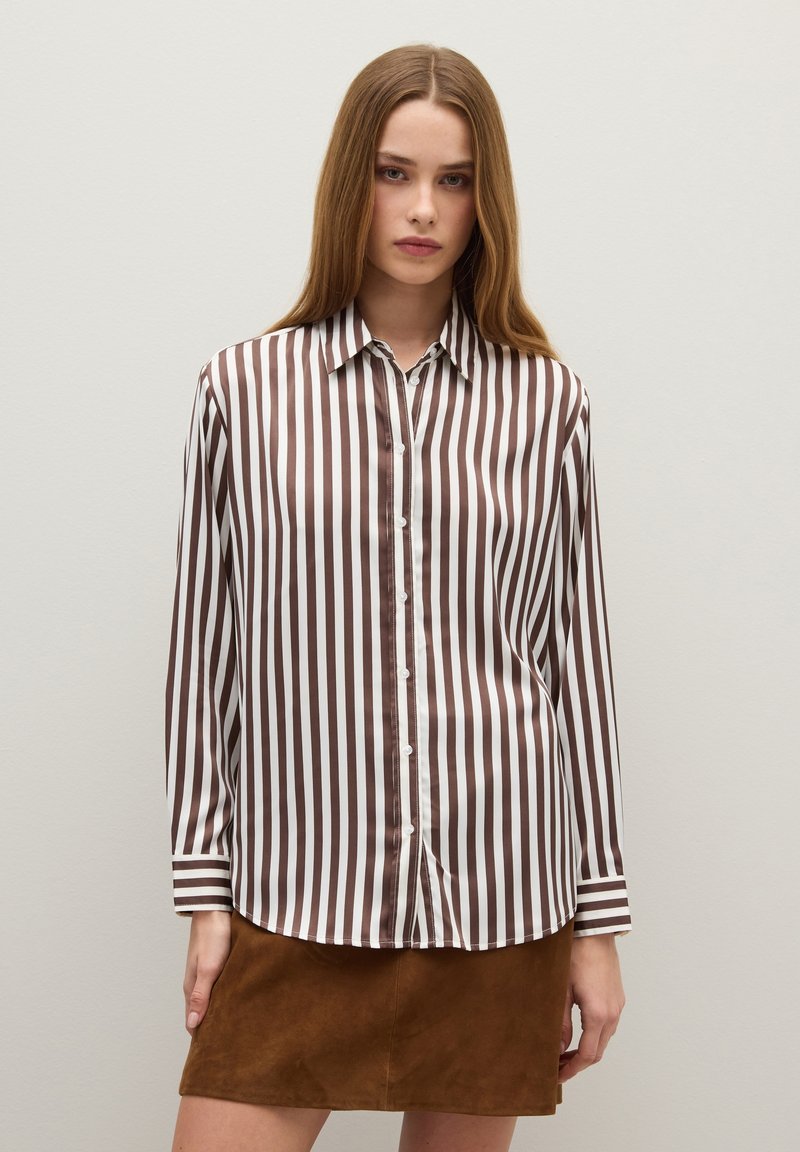 Young woman wearing a brown and white vertical striped button-up blouse and a brown suede mini skirt, standing against a plain wall.