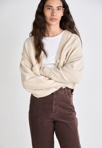 Beige gebreide cardigan met chunky textuur, grote knopen en voorzakken, gedragen over een witte shirt en bruine corduroy broek.