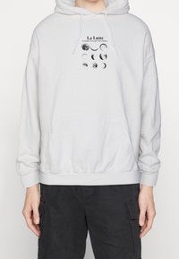 Ljusgrå hoodie med en frontficka. Har en svart grafisk tryck av månens faser och texten: "La Lune, En guide genom vår galax."
