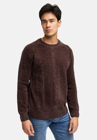 Jonge man in een gestructureerde donkerbruine gebreide trui en blauwe jeans, staand tegen een effen lichte achtergrond.