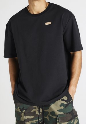 Sort bomulds T-shirt med rund hals, korte ærmer og et struktureret logo-patch på brystet, kombineret med camouflage cargo shorts.