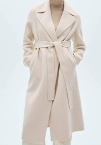 Abrigo de mezcla de lana de color beige claro, con cinturón, cuello con muescas, mangas largas y bolsillos laterales; presenta una textura suave y líneas limpias.