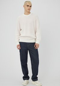 Pull en maille côtelée rose clair avec un col rond, associé à un jean droit bleu foncé et des baskets beiges. Texture douce, coupe décontractée.