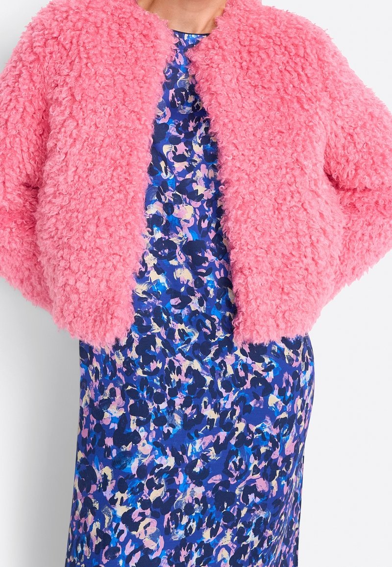 Vaaleanpunainen pörröinen cropped-takki, jossa on teksturoitu pinta, yhdistettynä siniseen kukkakuosisena mekkoon, jossa on erilaisia abstrakteja muotoja eri väreissä.