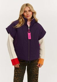Fioletowa kamizelka z ociepleniem, z wysokim kołnierzem, krótkimi rękawami i przednim zamkiem błyskawicznym. Nosi się ją na wielotkanej bluzie i spodniach w panterkę, z różową metką.