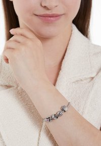 Zilveren armband met charms, waaronder een hart en complexe ontwerpen, tegen een gestructureerde, lichtgekleurde achtergrond.