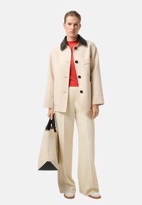 Beżowa kurtka z mieszanki wełny z czarnym kołnierzem, z guzikami i przednią kieszenią, w połączeniu z czerwoną bluzką, beżowymi spodniami i pasującą torbą typu tote.