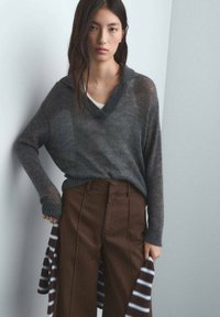 Pull gris surdimensionné avec un col en V et des manches longues, associé à un pantalon marron taille haute. Une chemise rayée dépasse sous le pull.