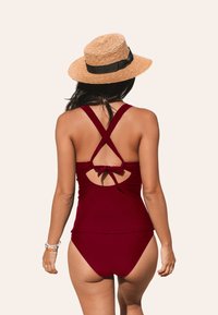 Bañador burdeos con un diseño de espalda cruzada, adornado con un lazo y detalle de espalda descubierta, combinado con un sombrero de paja con cinta negra.