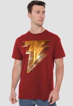 Man wearing a red T-shirt with a large shiny gold lightning bolt emblem, looking to his right against a plain background.