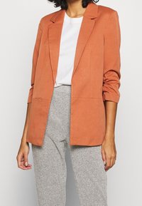 Marynarka w kolorze rdzy, o dopasowanym kroju, z wyciętymi klapami i dwoma przednimi kieszeniami. Nosi się ją na białą koszulę i wzorzyste szare spodnie.