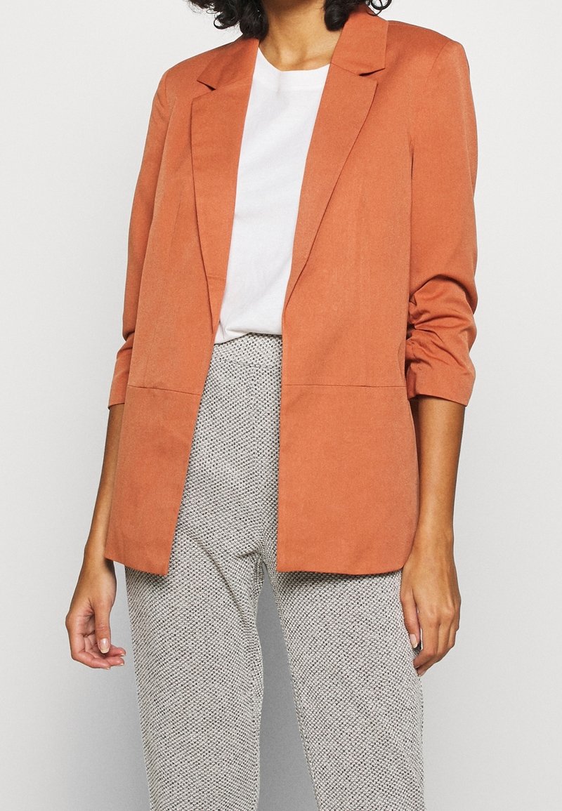 Marynarka w kolorze rdzy, o dopasowanym kroju, z wyciętymi klapami i dwoma przednimi kieszeniami. Nosi się ją na białą koszulę i wzorzyste szare spodnie.