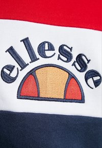 Červená, bílá a námořnická látka má vyšívané logo "ellesse" nad designem ve tvaru půlkruhu v žluté a červené barvě. Jemná textura, sportovní design.