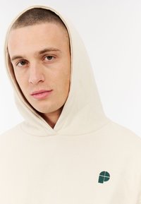 Beige hoodie met een trekkoord en een klein groen geborduurd logo op de borst. Zachte stof, ontspannen pasvorm en eenvoudig ontwerp.