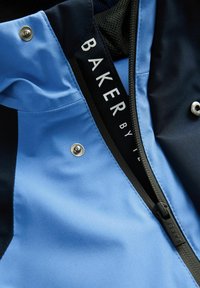 Lyst blå og marineblå jakke med en glat tekstur, der har en lynlås, trykknap og et mærke med teksten "BAKER by."
