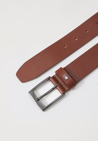 Ceinture en cuir brun avec une boucle en métal argenté rectangulaire, des accents cousus et un petit logo près de la boucle. Plusieurs trous pour ajustement.