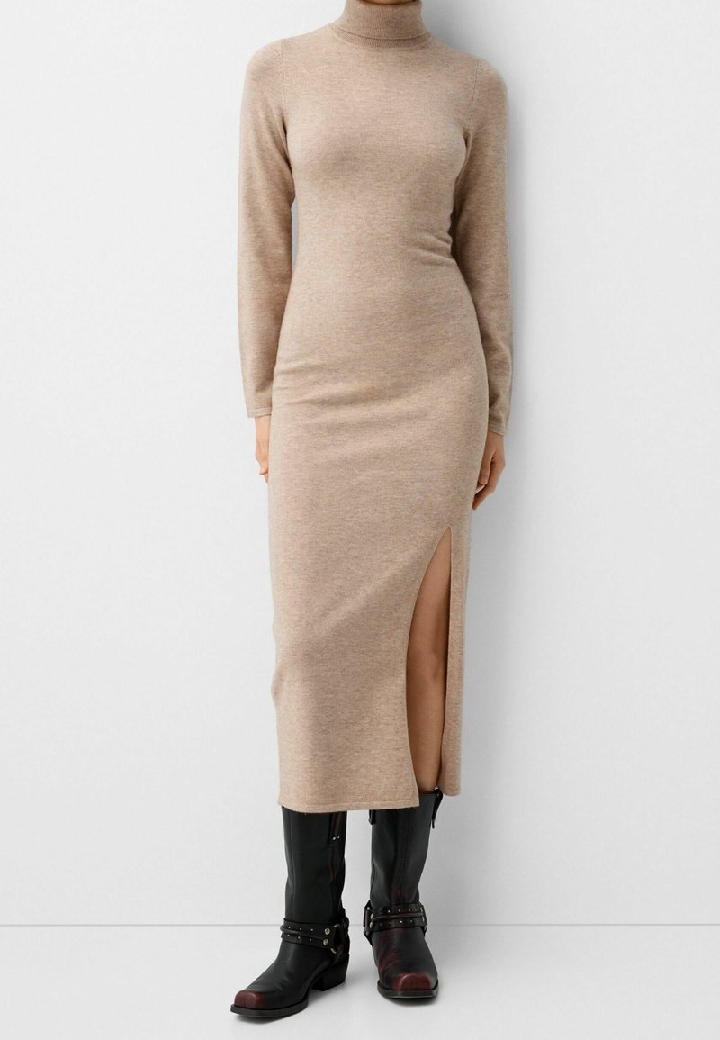Robe pull en maille beige à manches longues et col roulé, fendue sur le côté, portée avec des bottes en cuir noires mi-mollet munies de sangles et de boucles.