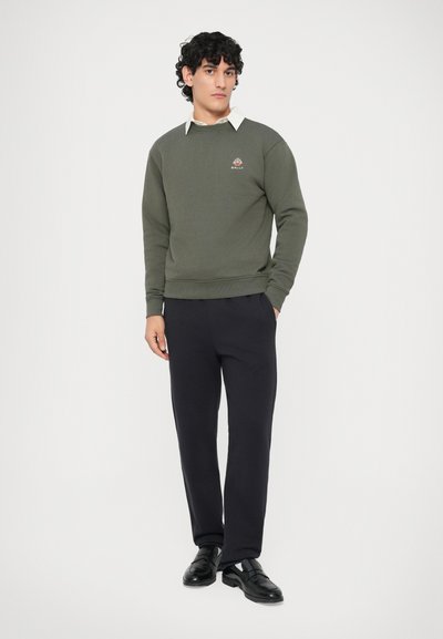 Mees, kellel on lokkis must juuksed, kannab oliivrohelist sweatshirti logoga, valget kaelusega särki, musti pükse ja musti ülikonnasaapaid, seisab rahulikult.