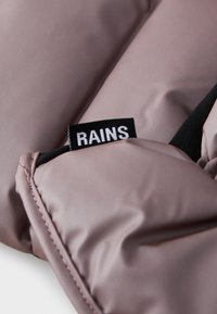 Hele lilla pehmeast tekstuurist polsterdatud jakk, millel on must brändi silt sõnaga "RAINS" ja kontrastne elastne materjal.