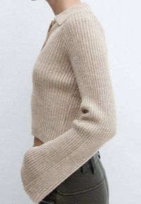 Pull en tricot côtelé beige coupé avec des manches évasées porté avec un pantalon noir taille haute sur un fond clair uni.