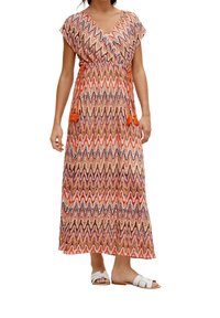 Femme portant une robe sans manches, longue jusqu'aux chevilles, à motifs zigzag multicolores avec une ceinture à franges orange et des sandales blanches à semelle plate.