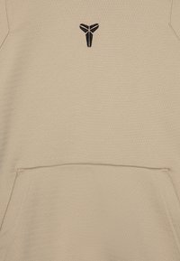 Beige hoodie i texturerat tyg. Har en framficka och en svart logotyp på bröstet. Enkel och funktionell design.