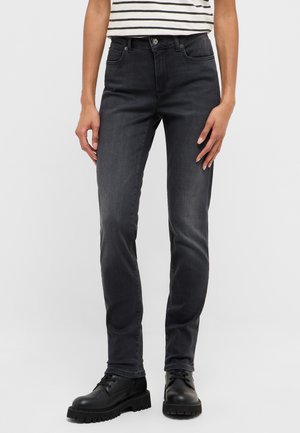 Person trägt ausgewaschene schwarze Jeans in schmaler Passform, schwarze Schnürstiefel und ein weiß-schwarz gestreiftes Hemd, steht vor einem einfarbigen Hintergrund.