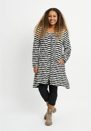 Gestreifter Langcardigan in Schwarz und Weiß mit einem Reißverschluss, Taschen und einem abgerundeten Saum, kombiniert mit schwarzen Leggings und tanfarbenen Schnürstiefeln.