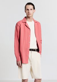 Chemise corail à boutons avec un col décontracté, portée sur un t-shirt blanc, associée à un short crème clair et une ceinture en cuir tressé noire.