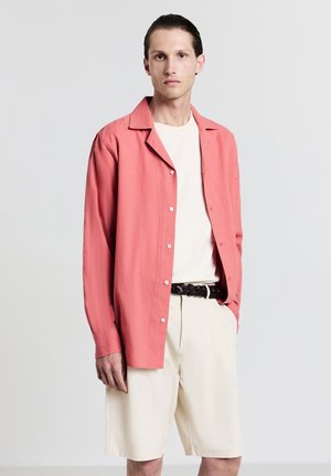 Chemise corail à boutons avec un col décontracté, portée sur un t-shirt blanc, associée à un short crème clair et une ceinture en cuir tressé noire.