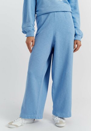 Hellblaue Jogginghose aus weichem Stoff mit weitem Bein und elastischem Bund. Kombiniert mit weißen Sportschuhen.