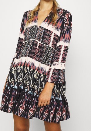 Kobieta nosząca sukienkę z długim rękawem sięgającą do kolan z wielokolorowymi plemiennymi i abstrakcyjnymi wzorami w kolorach czarnym, białym, różowym i niebieskim.