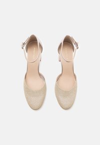 Para beżowych, błyszczących szpilek damskich na kostkę z zamkniętym noskiem i okrągłymi klamrami, widziana z góry na białym tle.