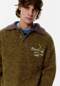 Neulottu oliivinvihreä paita, jossa on ruskea kaulus, nappilista ja kirjailtu teksti "Blooming with Care". Kankaan pinta on teksturoitu.