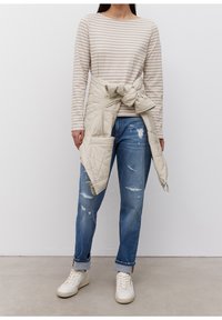 Beżowa i biała w paski koszula z długim rękawem, jasnobeżowa pikowana kamizelka związana w talii, zniszczone niebieskie dżinsy i białe trampki.