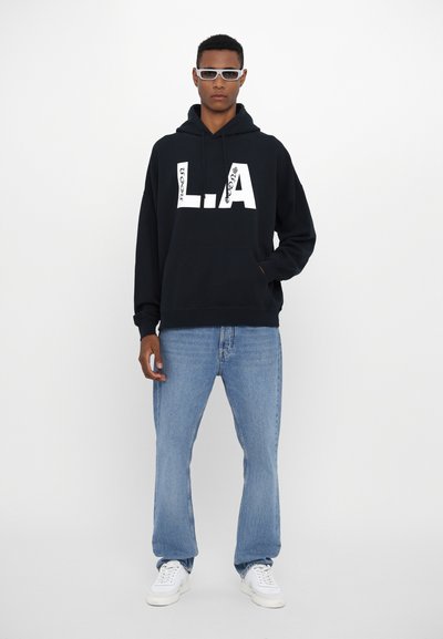 Black hoodie with large white "L.A." text and pattern, paired with light blue denim jeans and white sneakers. Model wears sunglasses.