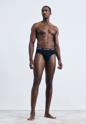 Izmos férfi mezítláb áll a fekete Björn Borg alsónadrágjában, egy sima világos háttér előtt, a kezei lazán az oldalán.