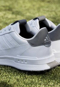 Sneakers bianchi con accenti grigi, materiale sintetico, lati perforati, collo imbottito e suola testurizzata con tre strisce sul lato.