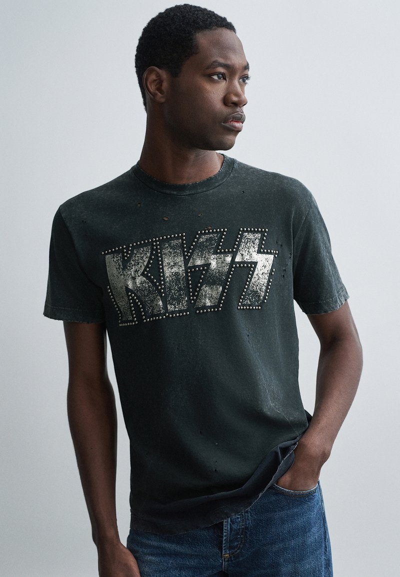 Camiseta negra desgastada con un logo metálico de "KISS" con tachuelas, mangas cortas y cuello redondo, combinada con jeans azules.