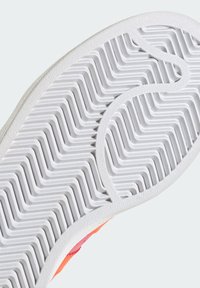 Sola de sapato com uma superfície de borracha branca texturizada, apresentando um padrão em zigzag e uma borda lisa, mostrando parcialmente uma parte superior em laranja e rosa.