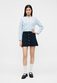 Νεαρή γυναίκα με ίσια μαύρα μαλλιά, φοράει ανοιχτό μπλε ζακέτα με σχέδια, σκούρο μπλε φούστα με κουμπιά, λευκές κάλτσες και μαύρα μοκασίνια, στέκεται με το χέρι στη μέση.