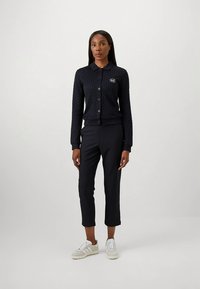 Tummansininen kuvioitu neuletakki napitettu, jossa on pieni logo, yhdistettynä mustiin räätälöityihin housuihin ja vaaleanharmaisiin tennareihin.