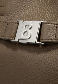 Lysbrunt tekstureret læder med en sølv metalspænde, der har et fremtrædende "B" logo, som viser en glat overflade og detaljeret syning.