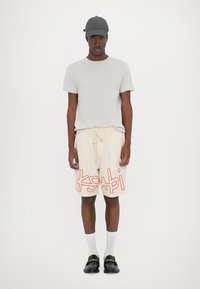 Grijze pet, grijs T-shirt, crèmekleurige shorts met rode "ksabi" tekst, witte sokken en zwarte instappers. De shorts hebben een gestructureerd patroon en een trekkoord.