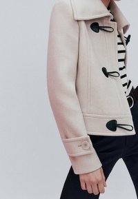 Jasno-beżowa kurtka z teksturowanej tkaniny, posiadająca kołnierz, czarne zapięcia na troczki oraz mankiety z guzikami. Noszona na paskowaną koszulę.